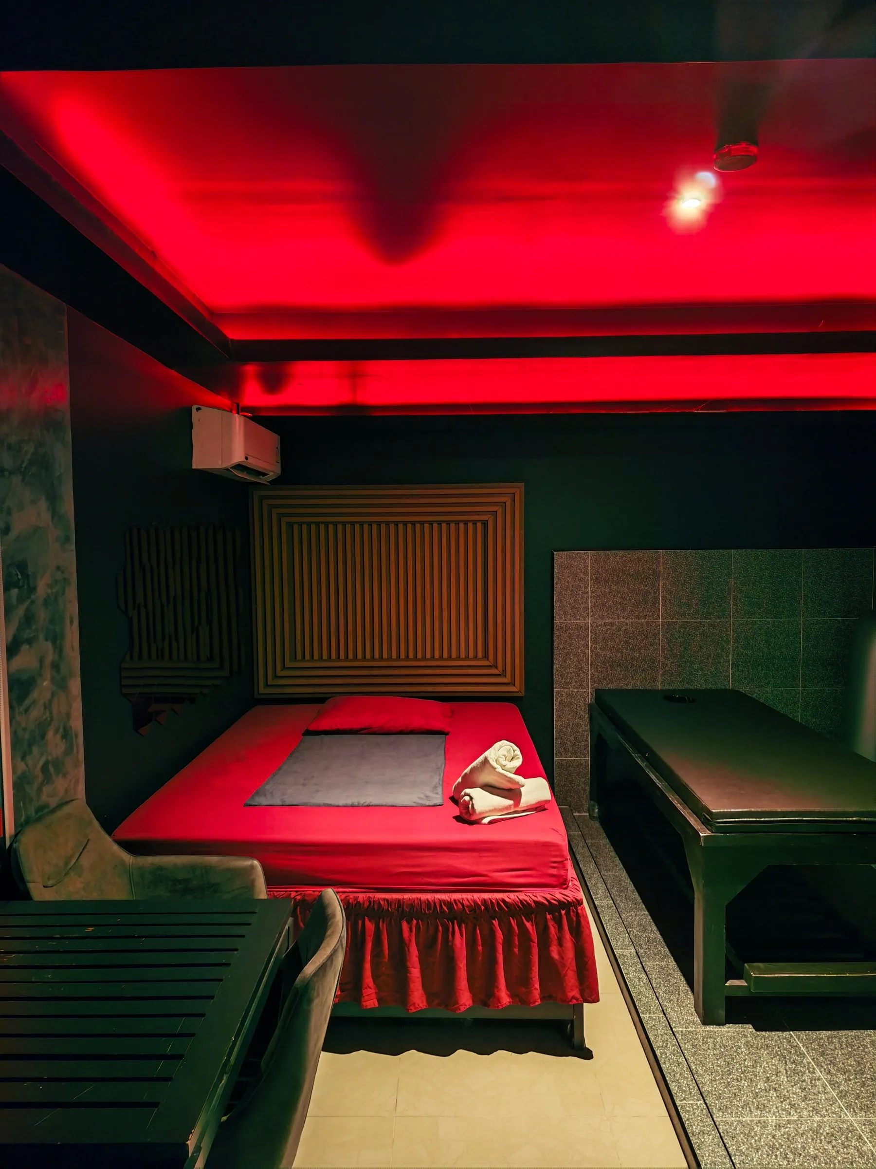 A massage room at Amor 888: a bed with red sheets and rolled towels beside a massage table, under a red-lit ceiling