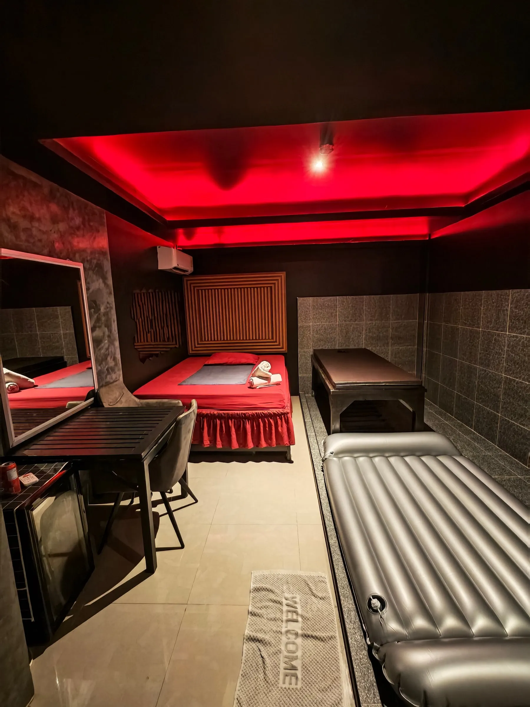 A private room at Amor 888: a bed with red sheets, a massage table, a table with two chairs and a mirror, and an air mattress on the tiled wet area