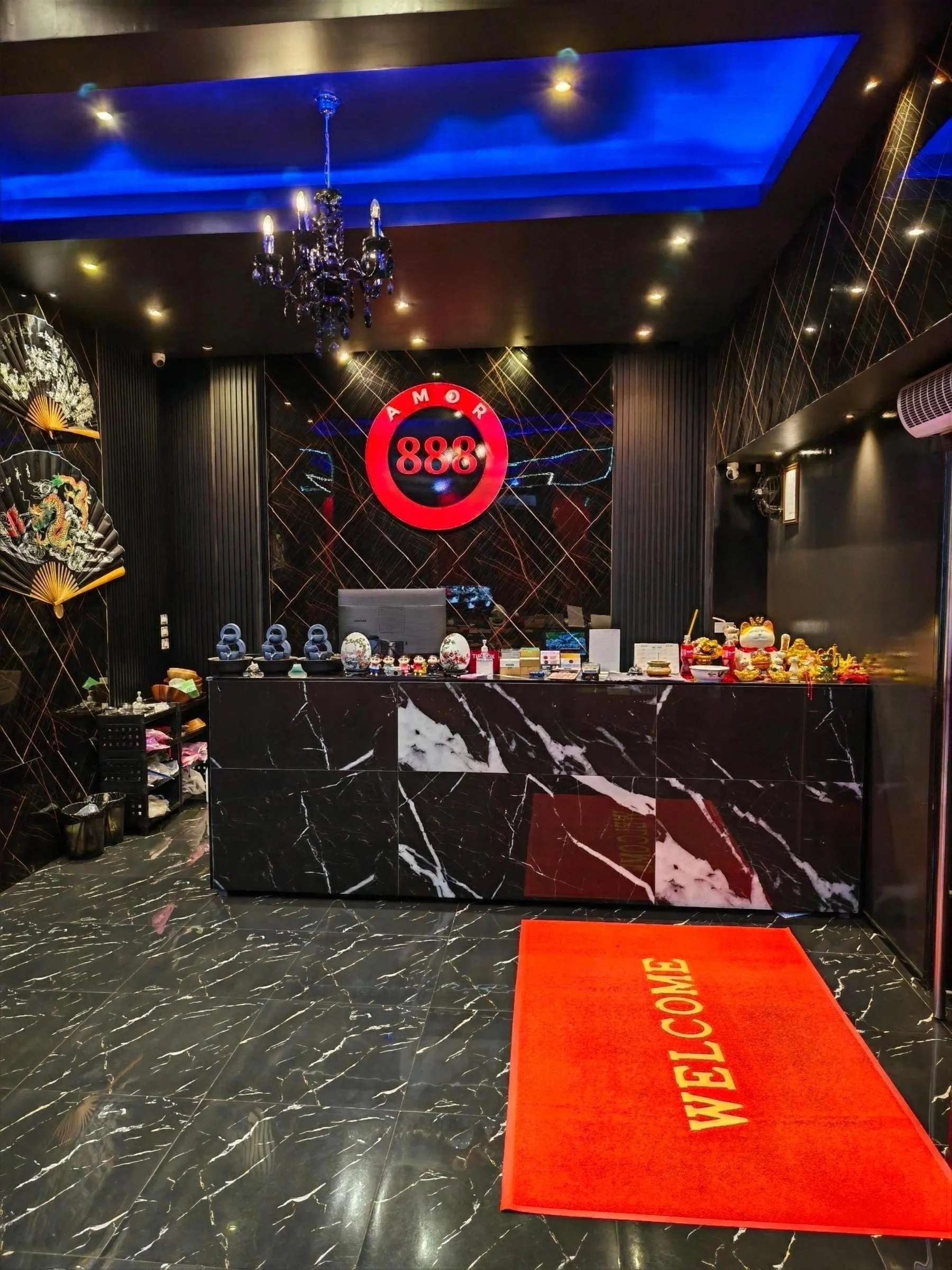 Reception at Amor 888: a black marble desk under the lit red 888 sign and a chandelier, with a red welcome mat on the floor